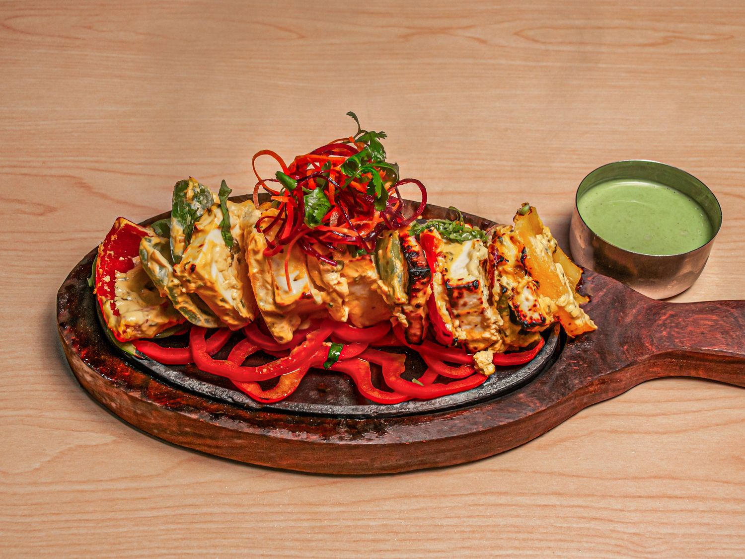 Afgani paneer tikka on a cast-iron sizzler over sliced red pepper, with a pot of green chutney