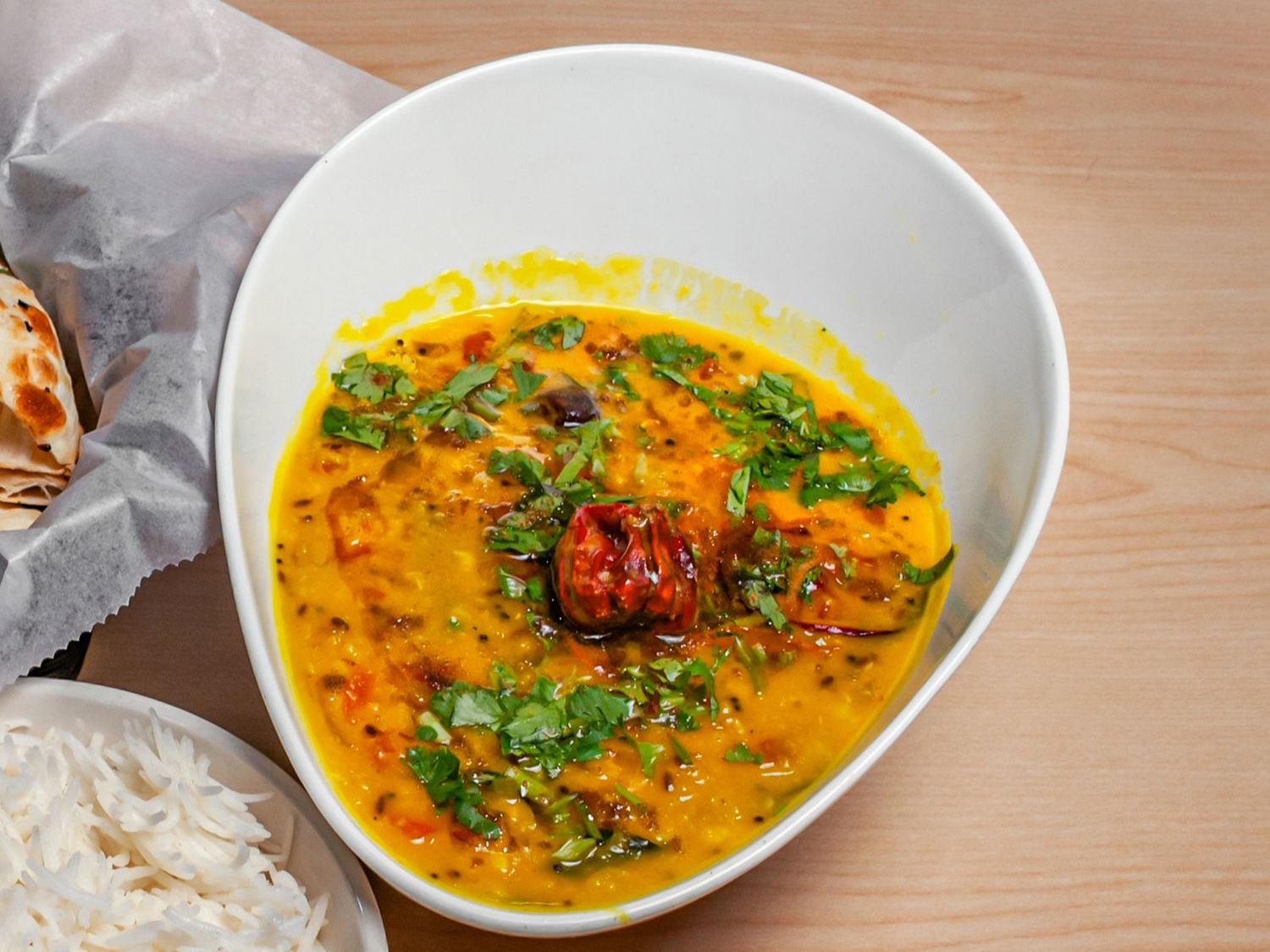 A bowl of BLR daal fry scattered with coriander and a whole red chilli, served with rice and bread