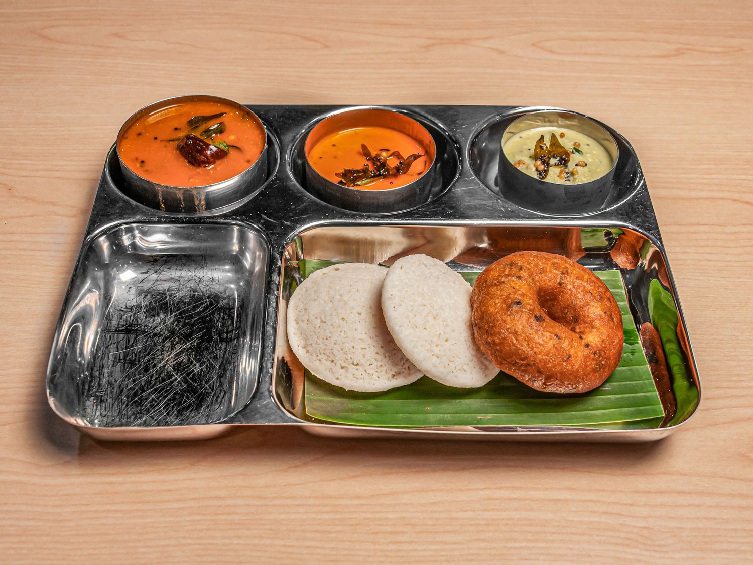 Two rice idli and a vada on a banana leaf, with sambar and two chutneys