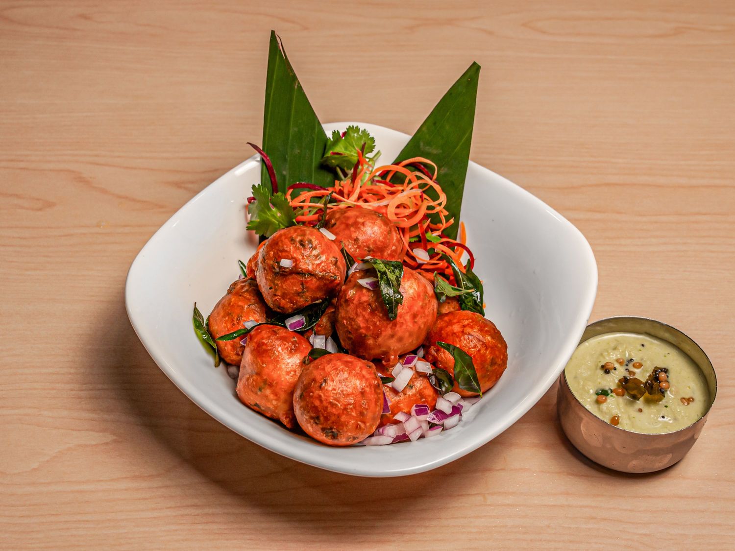 A bowl of Mangalore bajji with curry leaves and chopped onion, beside a pot of coconut chutney