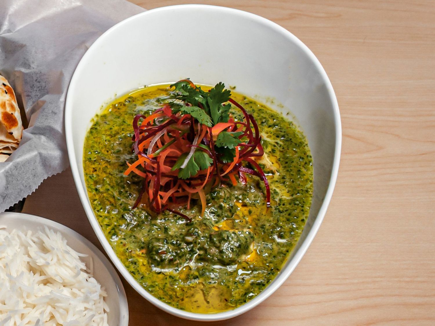 A bowl of palak paneer topped with slivered carrot and beetroot, with basmati rice and bread alongside