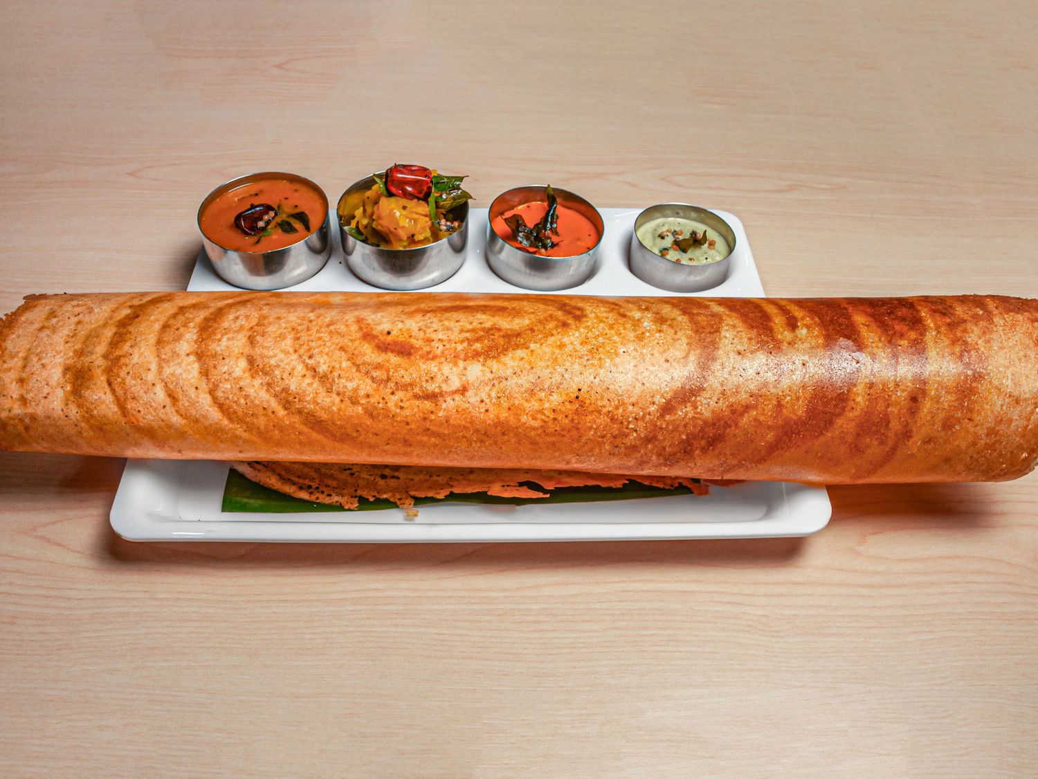 A long rolled paper dosa on a white plate, beside four katoris of sambar, potato masala and chutneys
